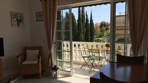 Une pièce comprenant une table à manger, des chaises, un fauteuil en osier, une petite table avec une lanterne, et une grande porte-fenêtre coulissante donnant sur un espace extérieur agrémenté d’une balustrade blanche et de verdure.