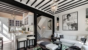 Un salon moderne avec un canapé blanc, un banc et une table basse. Un escalier mène à l’étage supérieur, et on y trouve également un coin cuisine avec des meubles blancs et un lustre.
