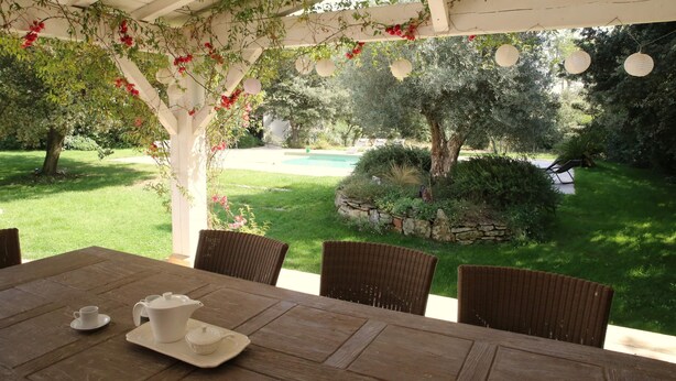 Un porche cubierto con una mesa lista para el té, rodeado de sillas de mimbre, con vista a una piscina y un jardín exuberante.