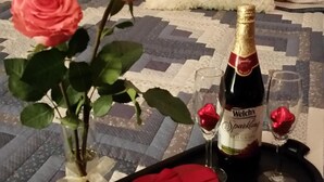 A bed with a quilted bedspread, a vase with pink roses, a bottle of champagne, two wine glasses, and a red napkin on a tray.