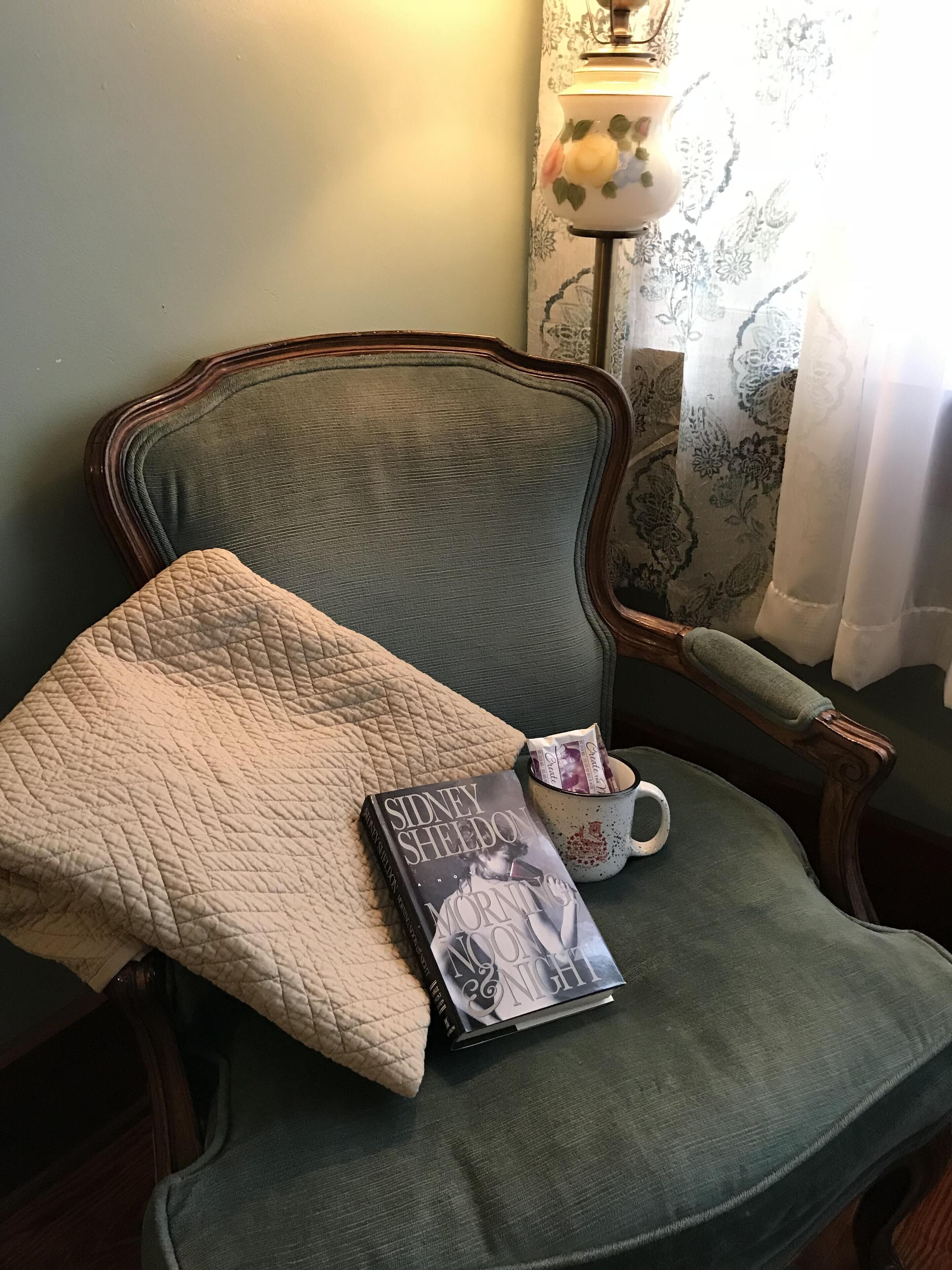 Ein grüner, gepolsterter Sessel mit einer gesteppten Kissenauflage, ein Buch mit dem Titel 'Mornings of the Moon' von Sidney Sheldon und eine Kaffeetasse mit floralem Dekor.