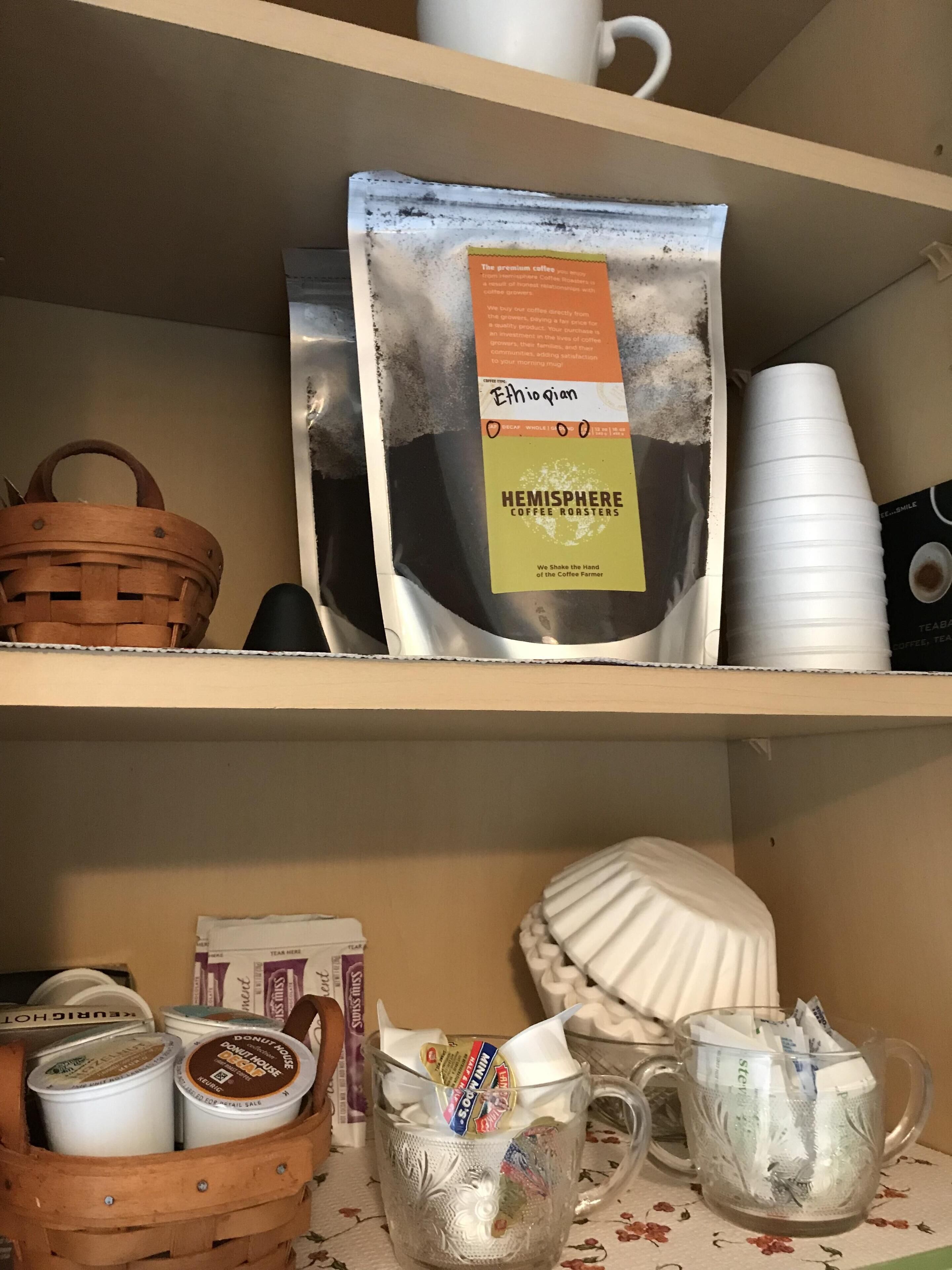 Hyllyllä on kahvipusseja, kertakäyttömukeja ja kori.
