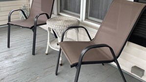A porch with two chairs and a small table, wooden floor, and a window with blinds.