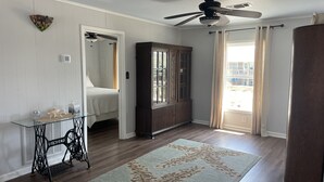 Une pièce comprenant un lit, une table en verre ornée d’un objet décoratif, un meuble en bois foncé, un ventilateur de plafond et une vue sur l’extérieur par une fenêtre.