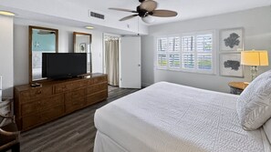 Ein Hotelzimmer mit Bett, Fernseher auf einer Holzkommode, Deckenventilator und Fenster mit Jalousien.