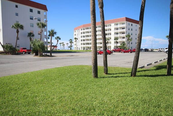 Un parking où sont garées plusieurs voitures rouges, avec des palmiers et un grand bâtiment blanc doté de balcons.