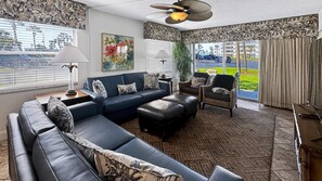Un salon moderne avec un canapé d’angle, une chaise longue, une table d’appoint avec une lampe, et une vue sur un parking grâce à de grandes fenêtres.