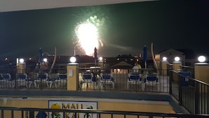 Vista nocturna de una zona costera con un espectáculo de fuegos artificiales, áreas de descanso y un cartel que dice 