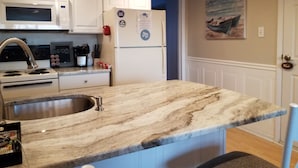 Una cocina con armarios blancos, encimera de mármol, pileta de acero inoxidable, microondas, refrigerador y un cuadro en la pared.