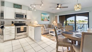 A modern kitchen with white cabinetry, a microwave, an oven, and a dining area with a table and chairs. There is a ceiling fan and a view of the outdoors through a large window.