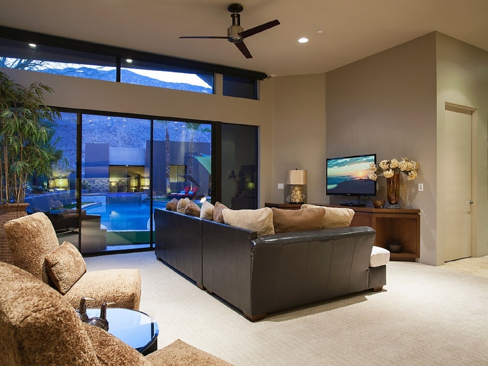 A modern living room with a large sectional sofa, a flat-screen TV, and a view of a swimming pool and mountains through large windows.