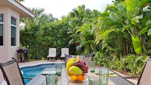 A poolside dining area with a table set for two, surrounded by lush tropical foliage.