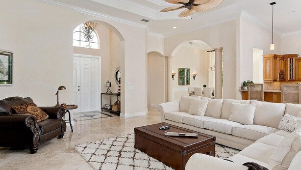 A spacious living room with a large sofa, a dark leather armchair, and a coffee table. There is a large window with a view of the outdoors, and a ceiling fan.