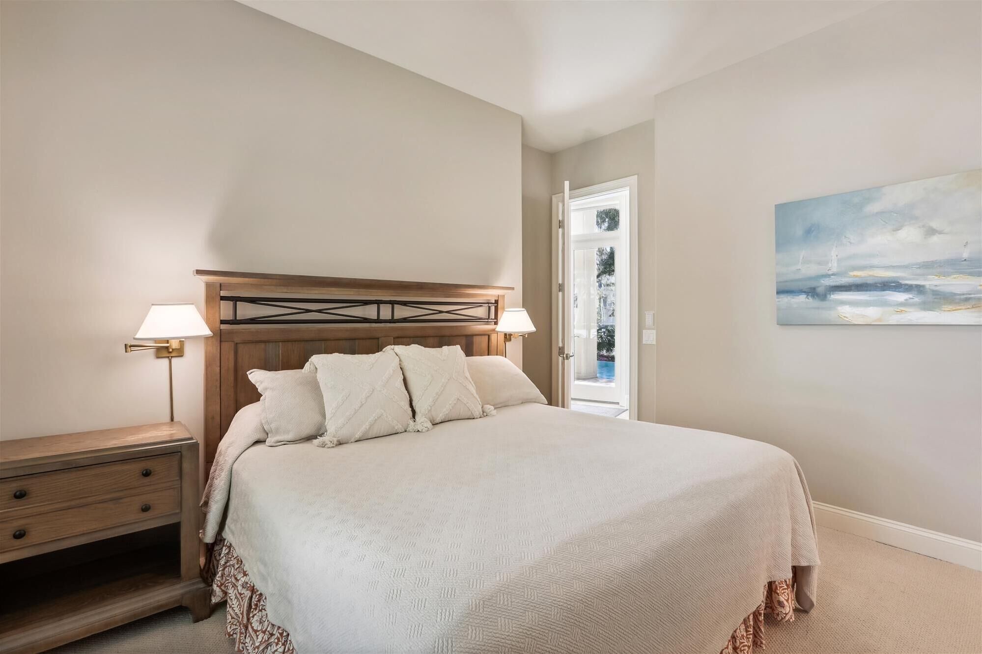 A bedroom with a wooden headboard, a bed with white bedding, a nightstand, a lamp, and a painting of a seascape.