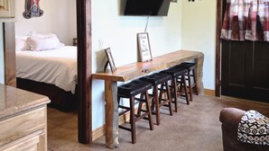 Ein Zimmer mit Esstisch aus Holz, einem an der Wand montierten Fernseher, einem Bett mit weißer Bettdecke und einer Tür zu einem weiteren Raum.