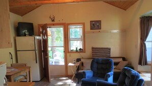 A cozy cabin interior with a wooden ceiling, a dining table, a blue armchair, a sofa, and a refrigerator. - A Cabin On The River<br>Three days minimum<br>Closed from October 15th to April 1 (Troy)