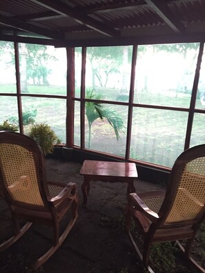 Ein Zimmer mit zwei Schaukelstühlen, einem kleinen Holztisch und großen Fenstern mit Blick auf einen Garten mit Bäumen und Pflanzen.
