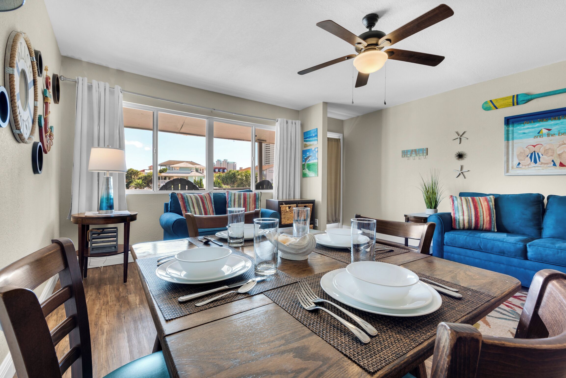 A dining area with a wooden table set for four, a ceiling fan, and a view of the outdoors.