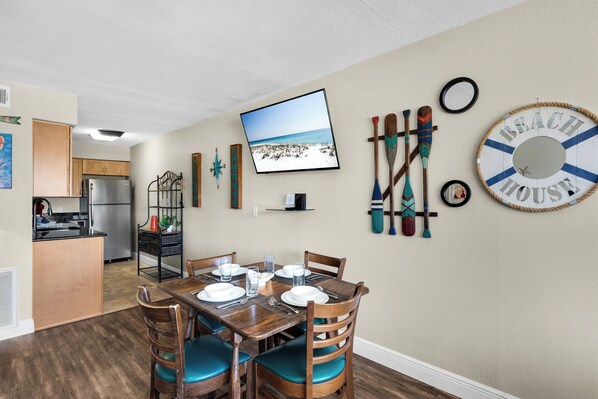 Un coin repas avec une table en bois dressée pour quatre personnes, une cuisine en arrière-plan et un téléviseur fixé au mur qui diffuse une scène de plage.