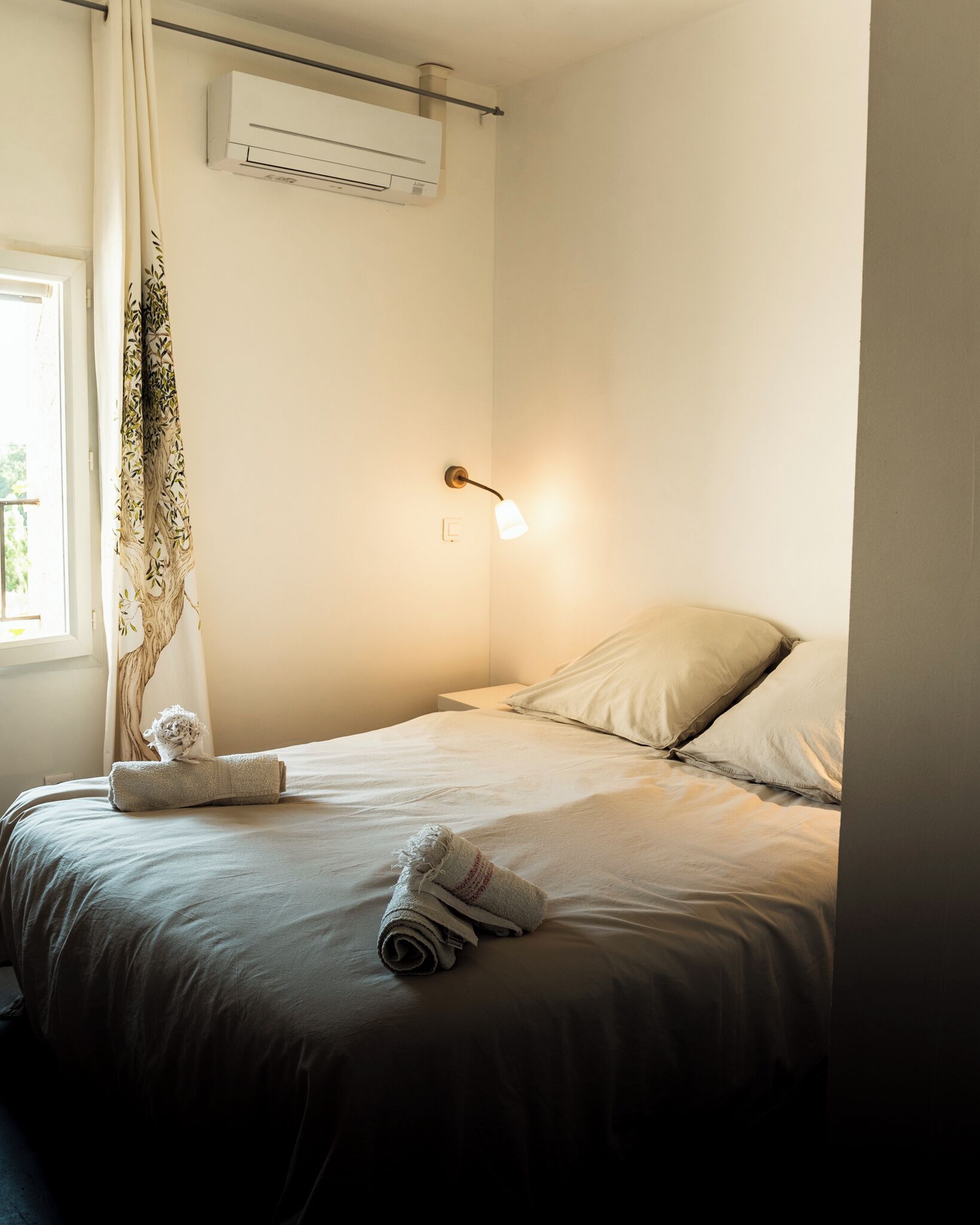 Un lit bien fait, avec une serviette roulée et un petit objet décoratif, un climatiseur fixé au mur et une lampe de chevet.