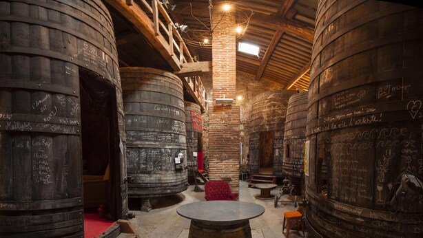 Un intérieur rustique agrémenté de grandes cuves en bois, d’une table ronde centrale et d’un escalier menant à un niveau supérieur.