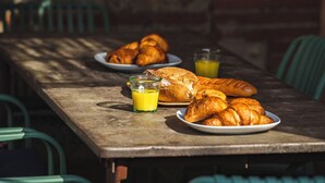Une table en bois, sur laquelle sont disposées des assiettes de croissants et un verre de jus d’orange, dans un coin repas agrémenté de chaises vertes.