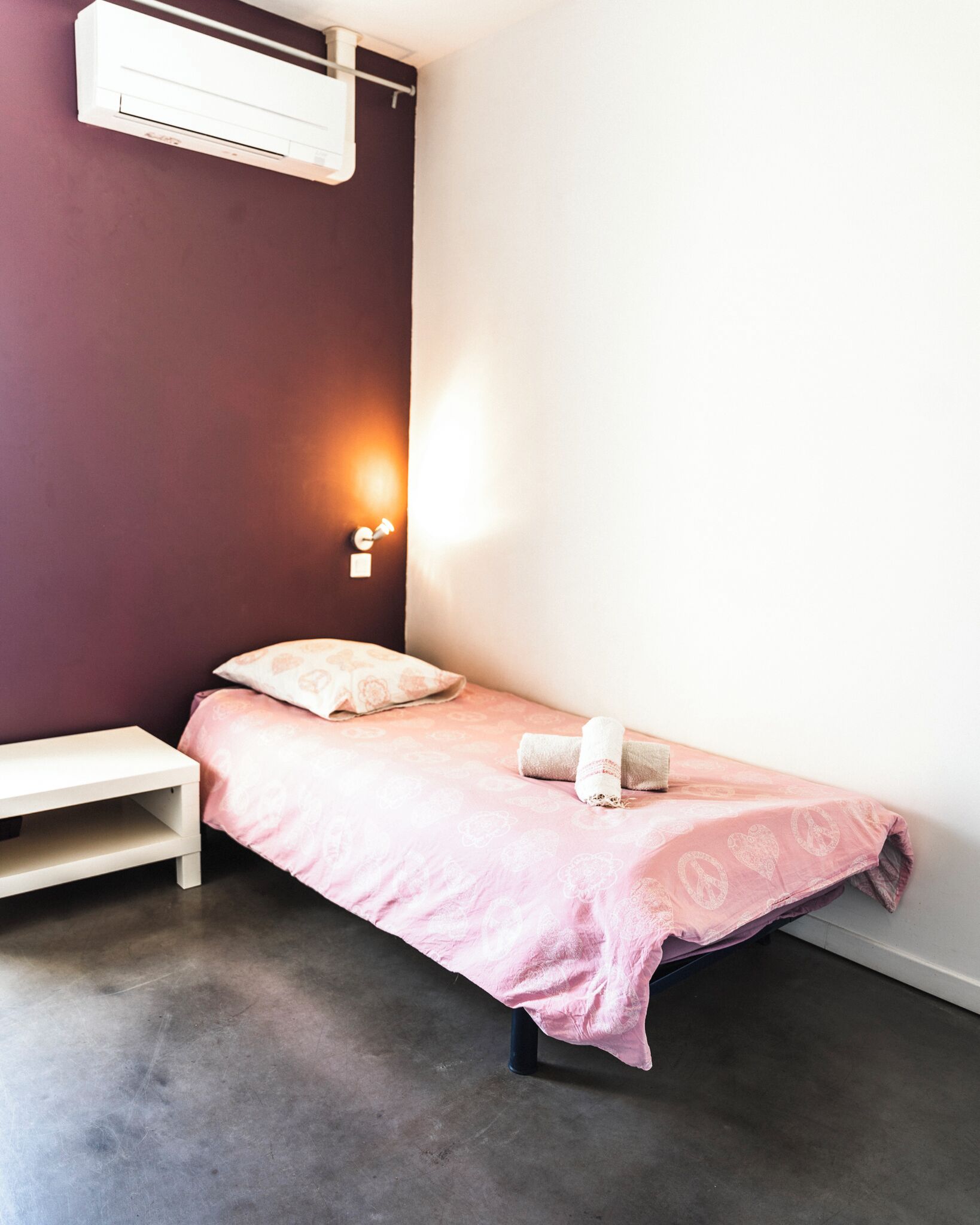 A small, single-bed room with a pink bedspread, a white bedside table, and a wall-mounted air conditioner.