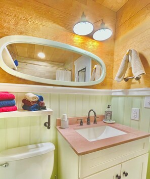 A bathroom with a wooden ceiling, a large oval mirror, a sink with a pink countertop, a toilet, and a shelf with folded towels. - SWEET HILL COUNTRY CABIN in the PERFECT location;  Sleeps 4-5 (Dripping Springs)