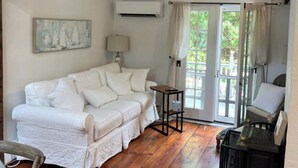 Une pièce chaleureuse avec un canapé blanc, un plancher en bois, un ventilateur de plafond et une vue sur l’extérieur, visible à travers une porte vitrée.