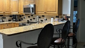 A modern kitchen with wooden cabinets, a stainless steel microwave, a granite countertop, and two bar stools.