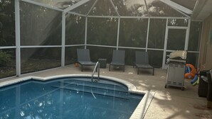 A covered outdoor swimming pool with a glass enclosure, lounge chairs, and a small storage cart.
