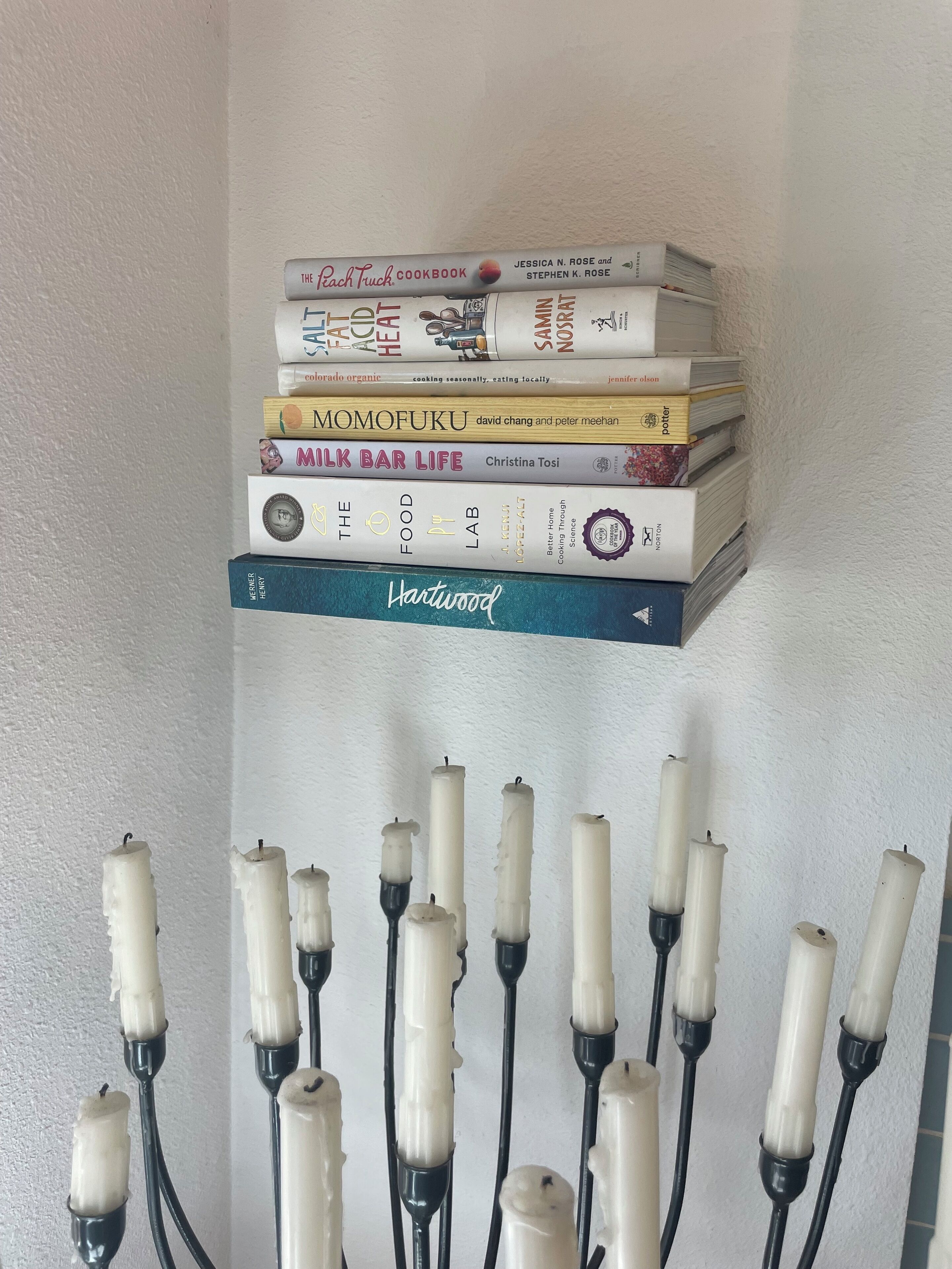 A stack of cookbooks on a shelf with candles below.