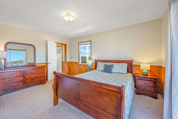 A bedroom with a wooden bed, a dresser, a nightstand, a mirror, and a window with a view. - Oceanfront Dog Friendly Home w/ Hot Tub, Game/Media Rooms, and Champagne Views! (Waves)