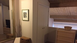 Een slaapkamer met een grote spiegel, een kledingkast, een klein tafeltje met een mand, en een raam met zonwering.