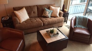 Een woonkamer met een bruine bank, twee fauteuils, een salontafel met decoratieve objecten, en uitzicht op het buitenbalkon.