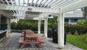 A covered patio area with a wooden picnic table, a barbecue grill, and a white pergola structure.