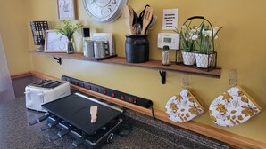A kitchen with a toaster, a clock, shelves with kitchen utensils, a thermostat, and a table with a cutting board.