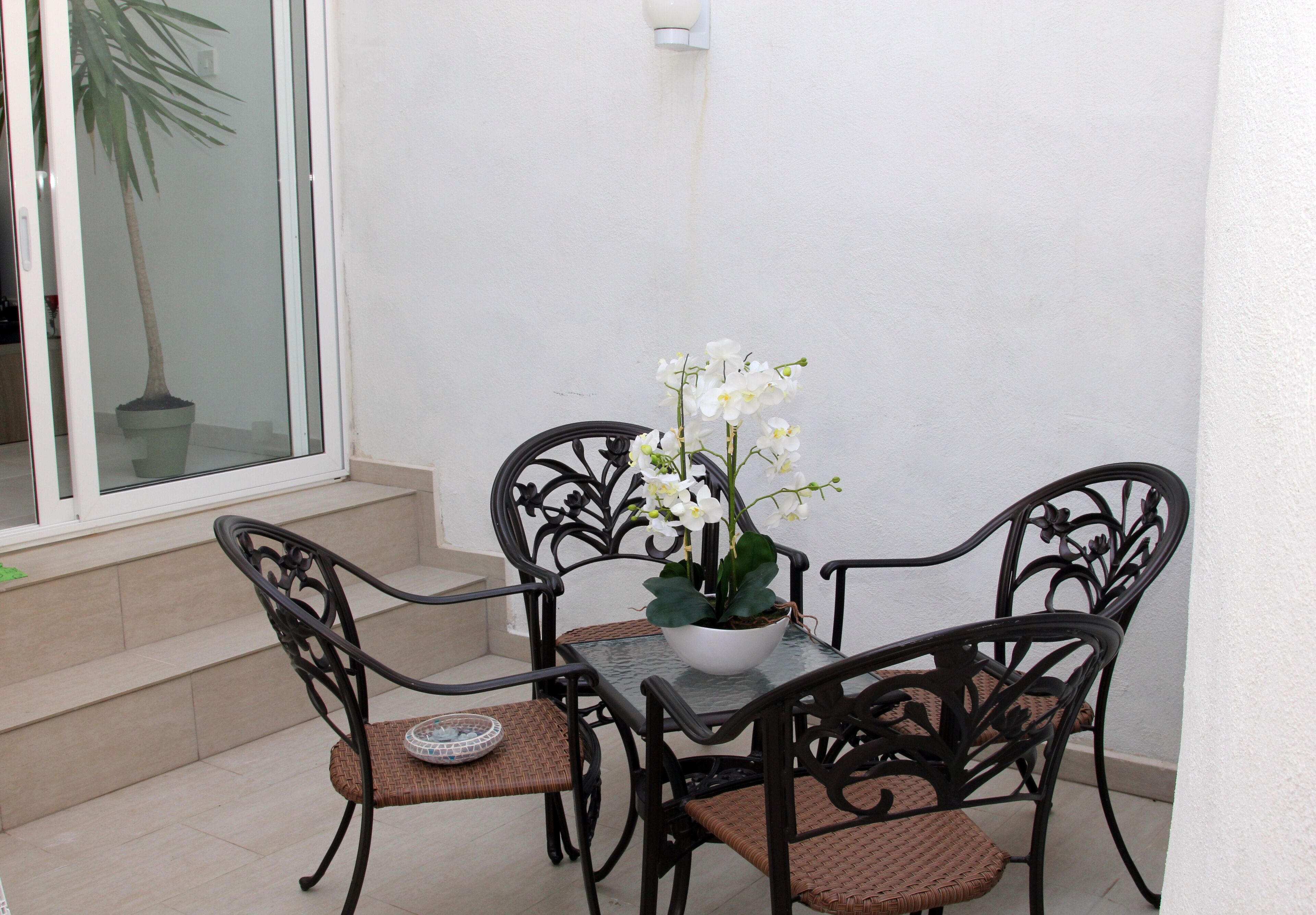 A small outdoor patio with a table and four chairs, a potted plant, and a view of a building through a glass door.