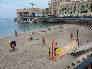 Uma praia com pessoas se banhando e tomando sol, uma torre de salva-vidas e edifícios ao fundo.