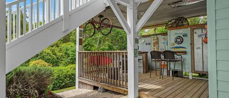 A covered outdoor area with a wooden deck, a bicycle hanging from the ceiling, and a small bar area with chairs and tables.