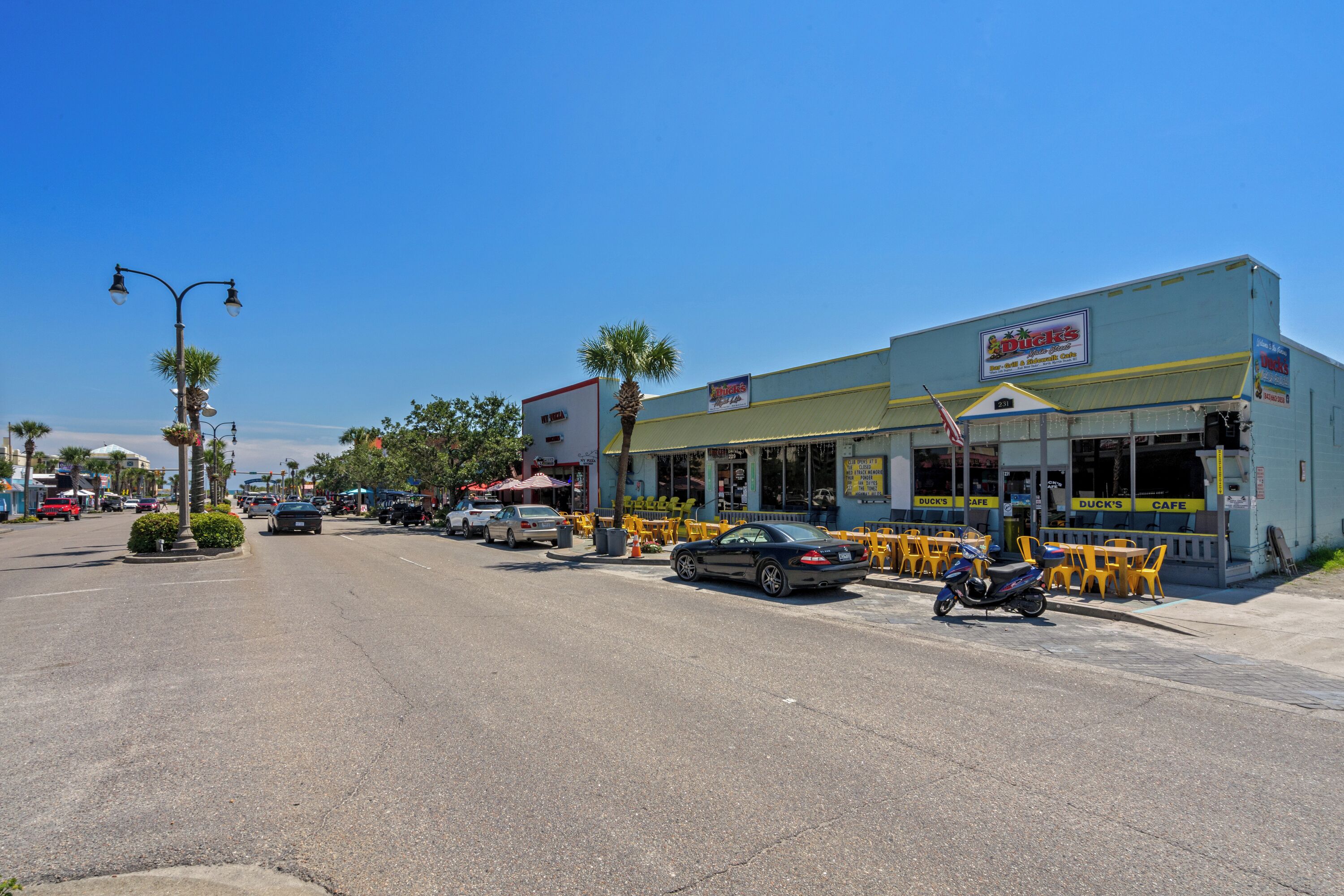 Vista de la calle con un edificio que dice “Duck's Cafe”, con mesas al aire libre y una motocicleta estacionada.