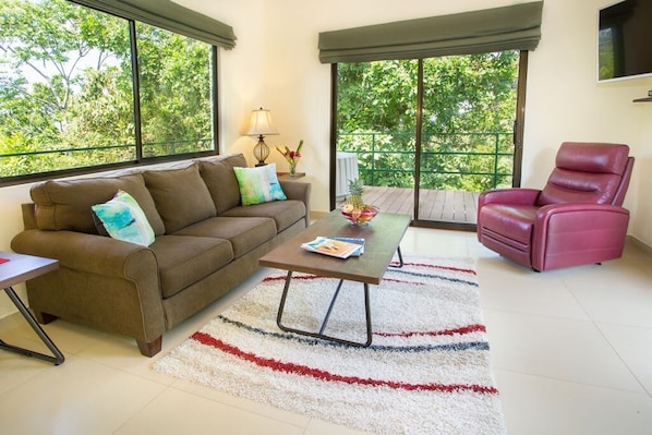 A living room with a brown sofa, a red chair, a coffee table with magazines, and a view of greenery through large windows.