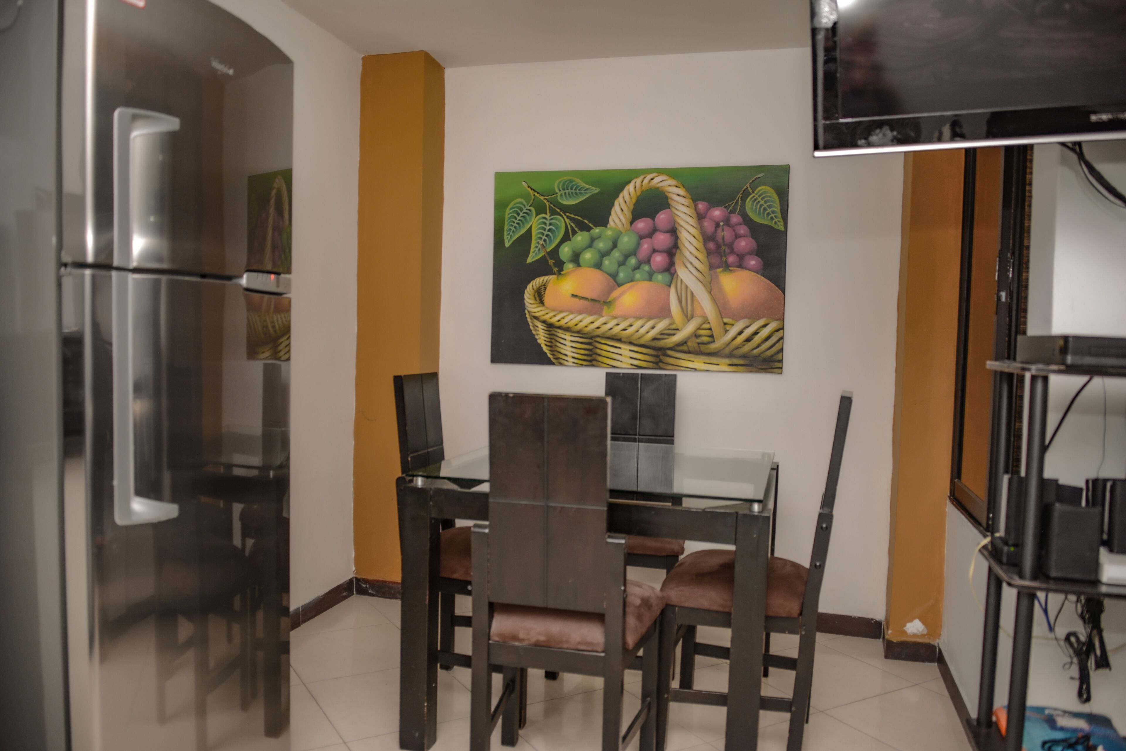 A dining area with a glass table and chairs, a refrigerator, a television mounted on the wall, and a painting of fruit.
