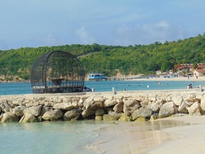 Paisaje costero con un rompeolas rocoso, agua azul cristalina y una playa con gente.