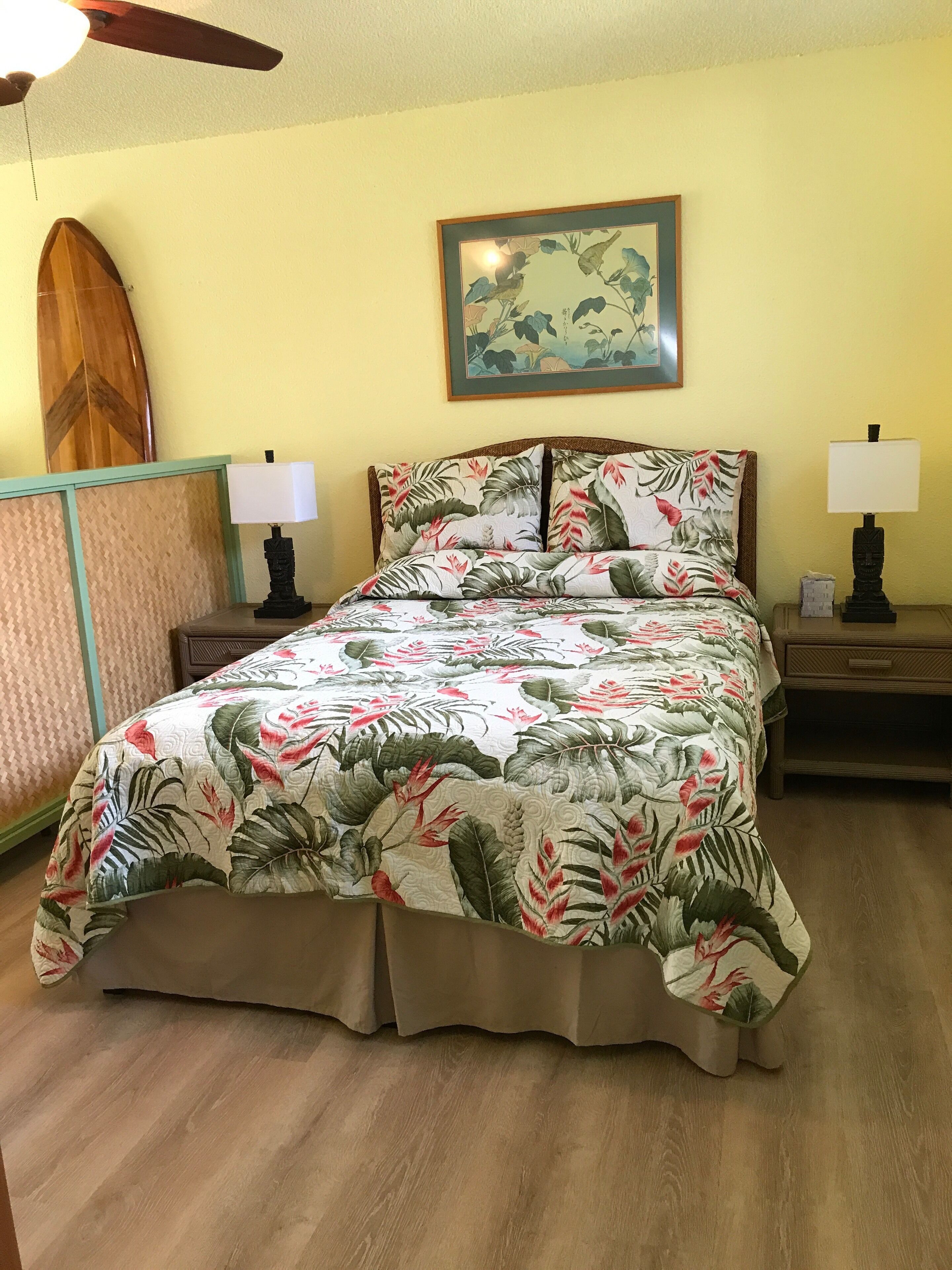 A bed with a floral bedspread, two bedside tables with lamps, a ceiling fan, a surfboard, and a framed picture on the wall.