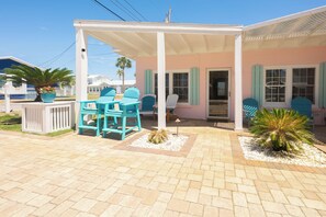 A covered outdoor seating area with chairs and a table, a building with a pink facade and teal shutters, and a palm tree.