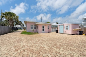 Uma casa móvel rosa com uma pequena varanda e uma garagem, cercada por um acesso pavimentado e uma cerca de madeira.