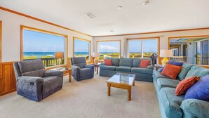 Ein geräumiges Wohnzimmer mit großen Fenstern und Meerblick, eine gemütliche Sitzecke mit Sofas und Sesseln sowie ein Couchtisch aus Holz.