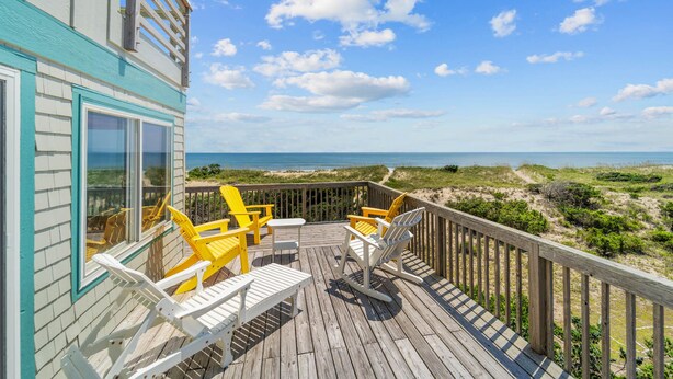 Eine Holzterrasse mit Gartenmöbeln, Blick auf den Strand und ein Gebäude mit blau-weißer Fassade.