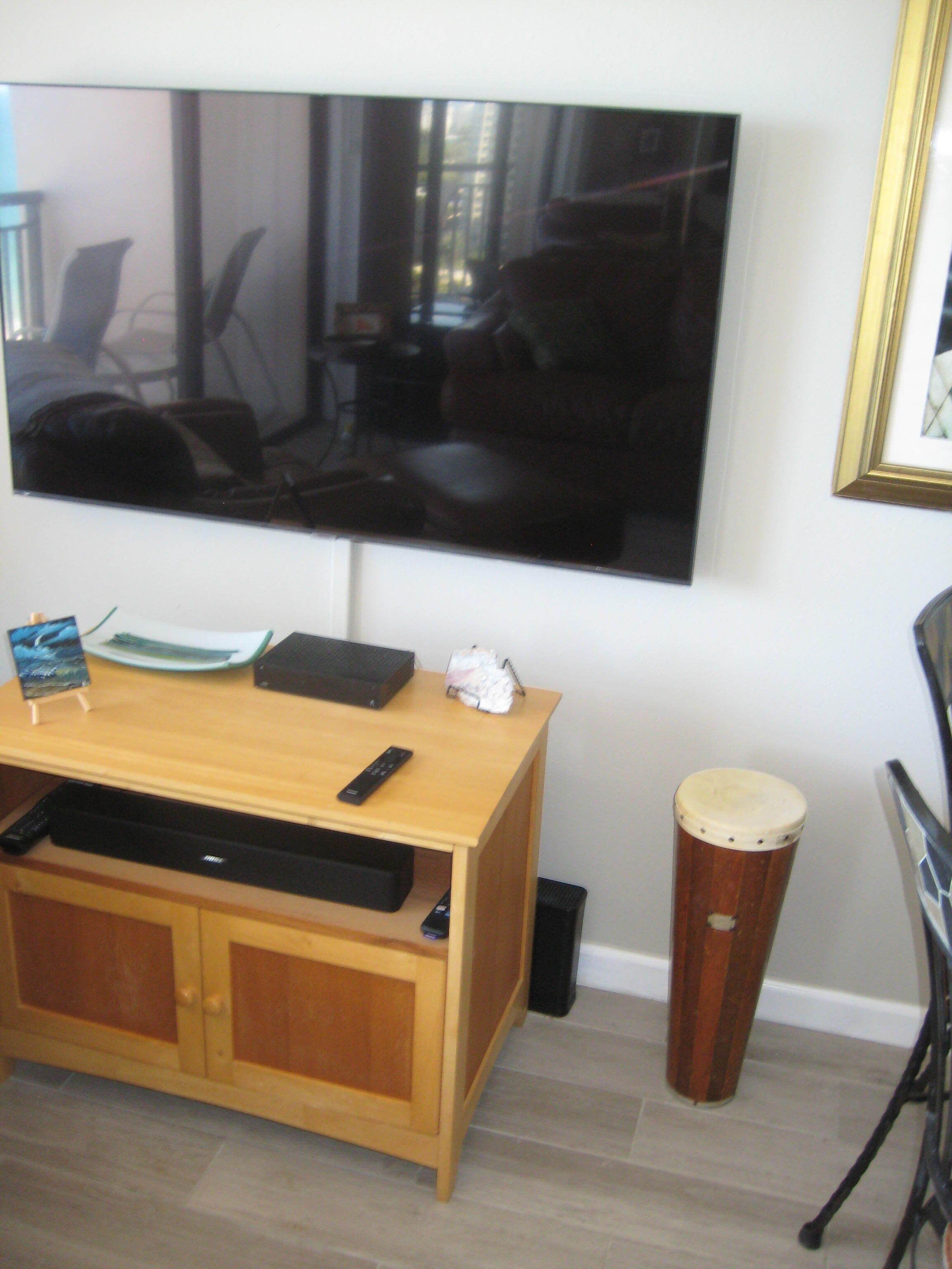 A living room with a flat-screen TV mounted on the wall, a wooden coffee table with remote controls, a trash can, and a drum.